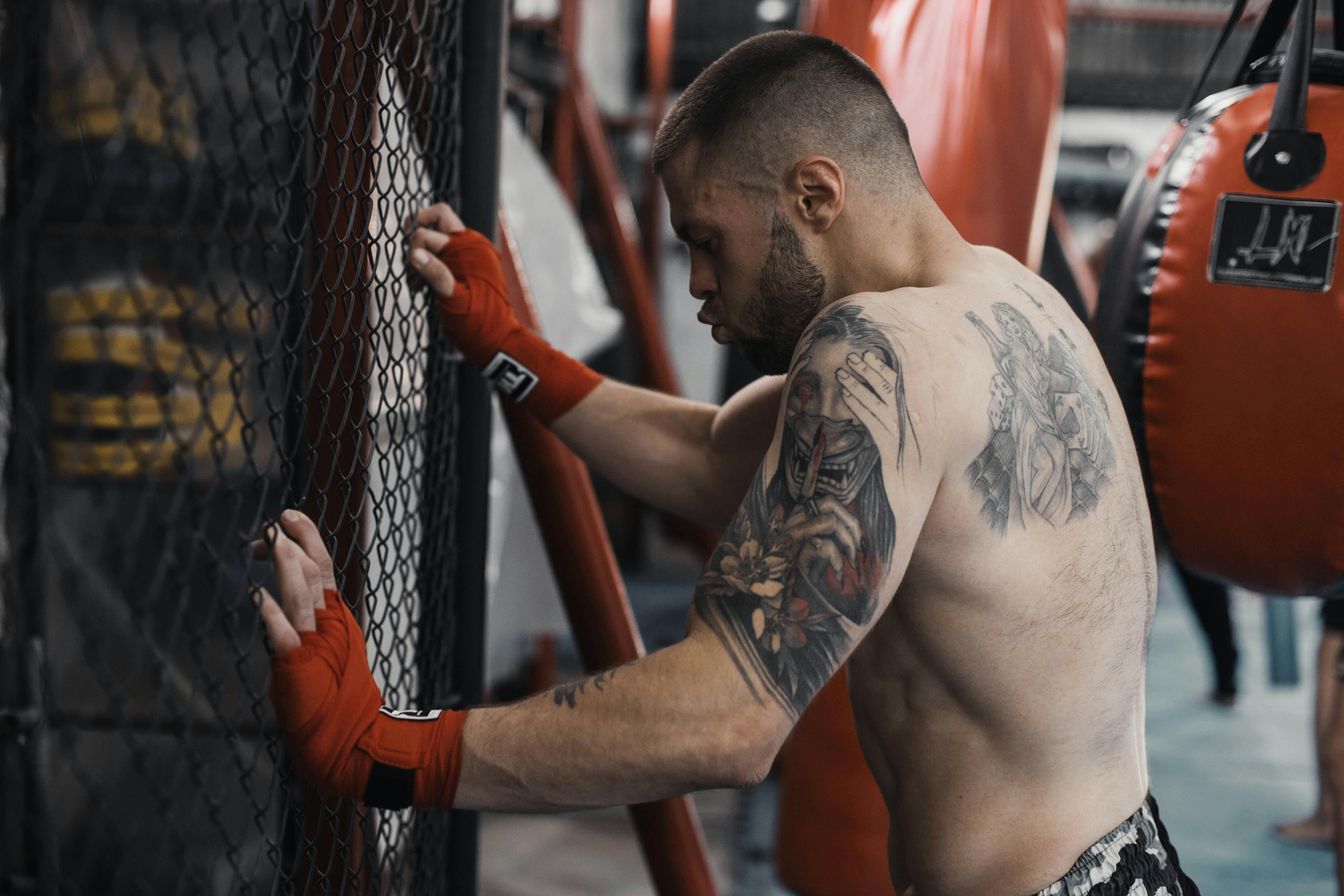A tattooed boxer taking a break while leaning against a gym fence with red boxing gloves.